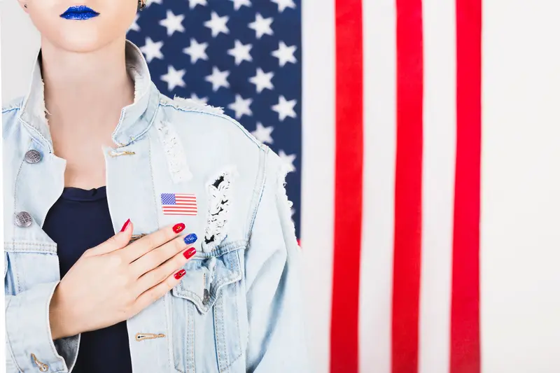 Eleições americanas - Mulher estadunidense com unhas pintadas nas cores da bandeira, bandeira dos EUA estampada na jaqueta e ao fundo bandeira dos EUA na vertical. 