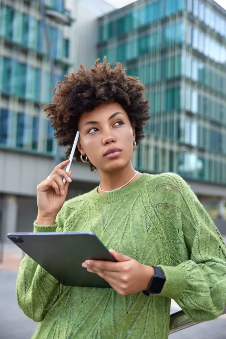 Muher negra vestindo uma blusa verde com seu tablet em frente a um prédio empresarial pensando sobre o futuro da educação executiva. 