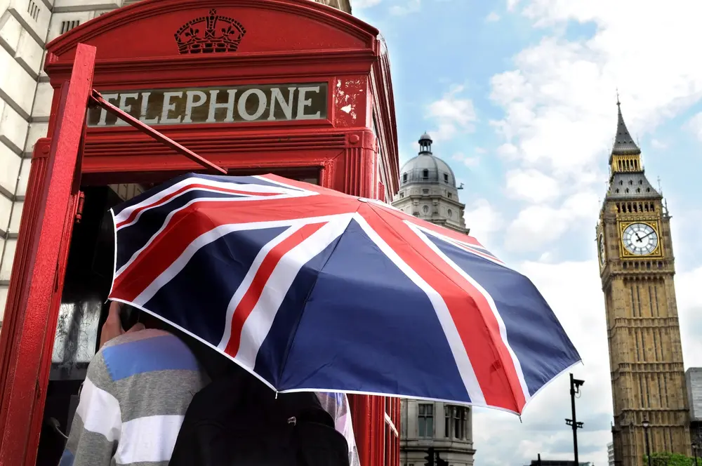 Pessoa aprendendo inglês executivo, em Londres, na foto com guarda-chuva com a bandeira britânica estampada, a cabine telefone inglesa vermelha e no fundo o big ben.