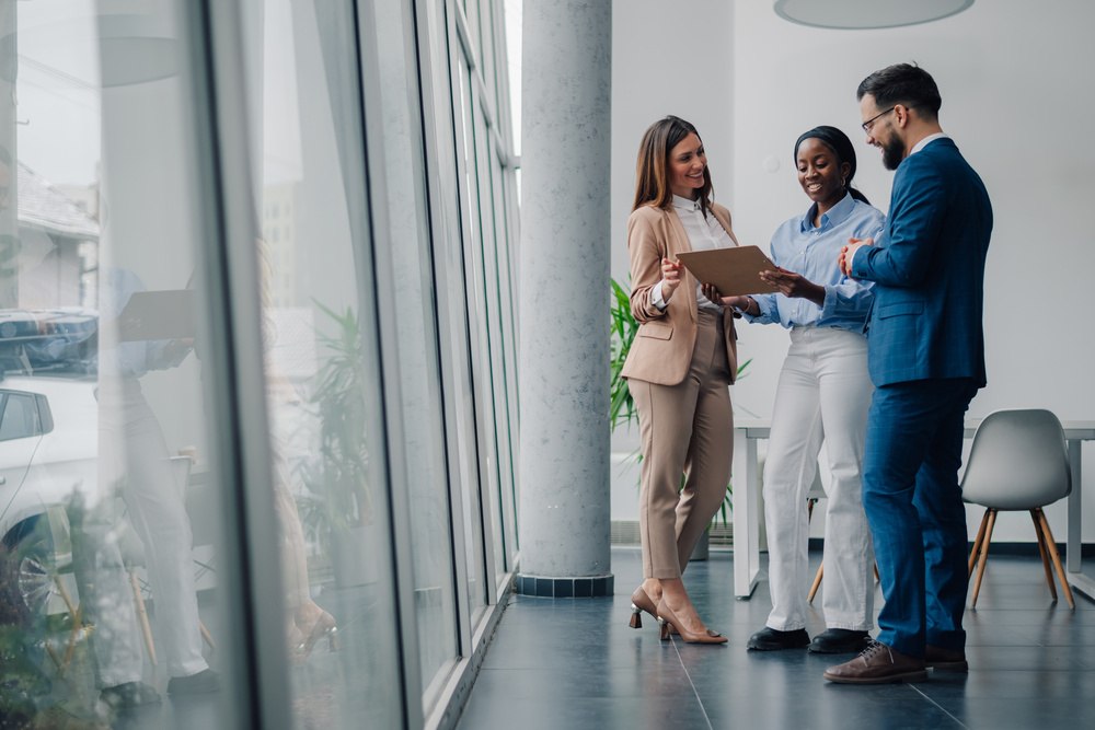 Três profissionais de negócios discutindo e analisando documentos em uma reunião de trabalho moderna, com grande janela de vidro ao fundo, transmitindo produtividade.