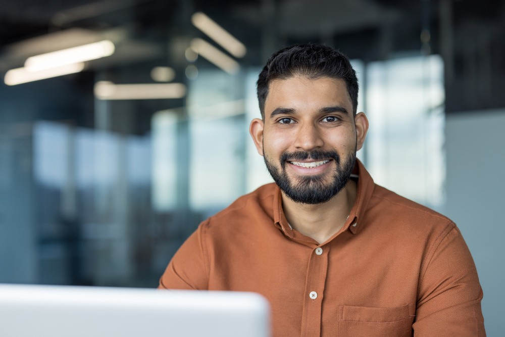 Profissional sorrindo em ambiente corporativo moderno, representando a alta dire&ccedil;&atilde;o em reuni&atilde;o estrat&eacute;gica diante do computador.